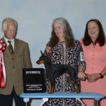 male judge holds red ribbon, woman poses with toy manchester terrier on table with third woman looking on