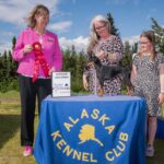 Show picture, judge is holding a group two ribbon & Jerri & Junior Chloe are posing with toy manchester terrier. The cloth over the table says 'Alaska Kennel Club'. Picture is outside & it is obviously windy.