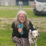 Jerri posing with Dalmatian & a new title ribbon, qualifying ribbon & a first place ribbon. Jerri is in front of an xpen & a sprinter van is in the background.