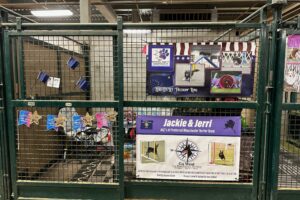 Decorated stall from 2023 AKC Agility nationals. One banner says 'Zero G's Let Freedom Ring' with many pictures of a Miniature American Shepherd. The other says 'Jerri & Jackie, AKC's #1 Preferred manchester Terrier Team' with a Logo for Go West Dogs (compass with manchester head in the middle) and pictures of manchester terrier doing agility.