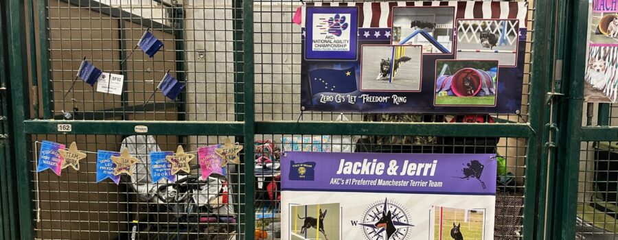 Decorated stall from 2023 AKC Agility nationals. One banner says 'Zero G's Let Freedom Ring' with many pictures of a Miniature American Shepherd. The other says 'Jerri & Jackie, AKC's #1 Preferred manchester Terrier Team' with a Logo for Go West Dogs (compass with manchester head in the middle) and pictures of manchester terrier doing agility.