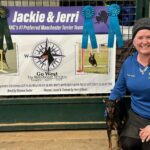 Jerri Posting with Jackie in front of Banner which says 'Jerri & Jackie, AKC's #1 Preferred manchester Terrier Team' with a Logo for Go West Dogs (compass with manchester head in the middle) and pictures of manchester terrier doing agility. Bred by Dianna Texter Owned, Loved & Trained by Jerri Gillean. GCHB CH PACH Bayside's Christmas Gift to Go West BN RA OA MXJ MXPE MXPB MJP3 PAX XF XFP T2B2P CA DCAT TKN